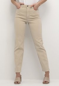 Pantaloni beige a vita alta con una vestibilità slim, dotati di tasche frontali, passanti per cintura e dettagli di cucitura discreti. Abbinati a tacchi aperti color nude.