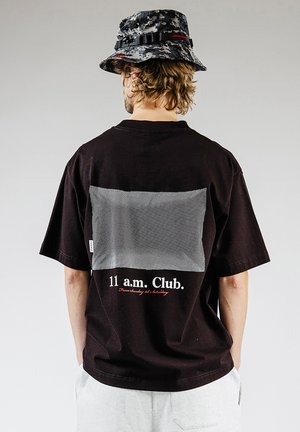 Osoba w czarnej koszulce z napisem "11 a.m. Club" i dużym szarym prostokątem na plecach, w połączeniu z kapeluszem typu bucket w kamuflażu i jasnymi szortami.