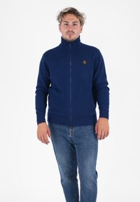 Maglione zip-up blu navale con colletto e polsini a coste, caratterizzato da un piccolo logo testurizzato sul petto. Indossato sopra dei jeans in denim.