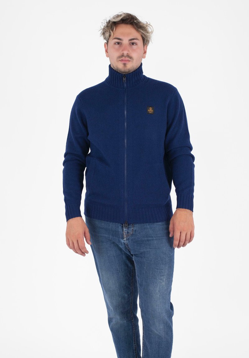 Maglione zip-up blu navale con colletto e polsini a coste, caratterizzato da un piccolo logo testurizzato sul petto. Indossato sopra dei jeans in denim.