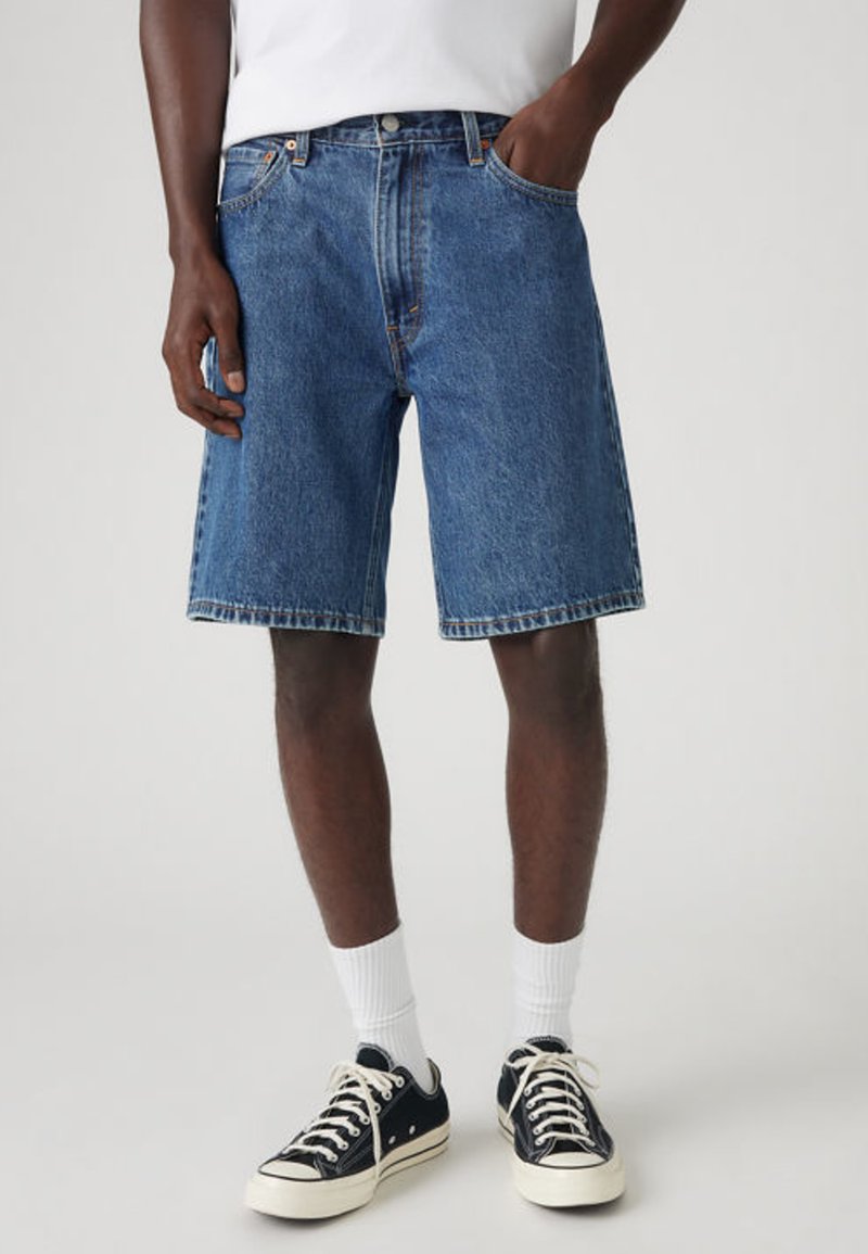 Homme debout portant un short en denim bleu, des chaussettes blanches, des baskets noires et une chemise blanche avec une main dans la poche.