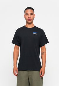 Fekete pamut póló kerek nyakkivágással, rövid ujjakkal és egy kék Nike logóval a bal mellen. Lazább fazonja és sima textúrája van.