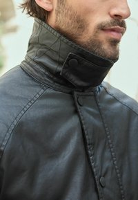 Sort jakke med høj krave, der har en glat tekstur, knaplukning og synlige stikninger på kraven.