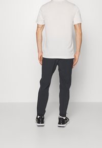 Męska dopasowana jasnoszara koszula z krótkim rękawem, w komplecie z ciemnoszarymi spodniami elastycznymi i czarnymi butami sportowymi, widziana od tyłu.