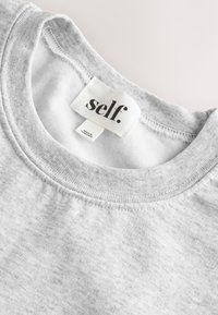 Šviesiai pilkas apvalia iškirpte džemperis, vidinėje apykaklės pusėje yra baltas lipdukas su užrašu „self.“ ir mažesnis lipdukas, kuriame parašyta „Pagaminta Hondūre.“