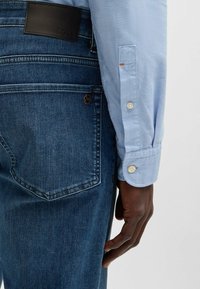 Ljusblå skjorta med uppvikt ärm, i kombination med mörkblå jeans som har en svart läderlogotyp och metalliska detaljer.