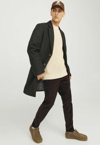 Un manteau en laine noir, un pull en tricot crème, un pantalon en velours côtelé marron, et des chaussures marron, avec une casquette marron arborant un logo, sur un fond neutre.