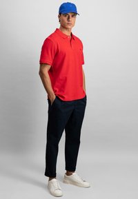 Rød polo-skjorte med krage, marineblå bukser og hvite sneakers. Blå cap gir fargekontrast. Enkel design uten synlige mønstre.