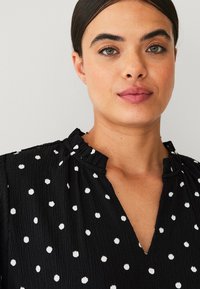 Black blouse with white polka dots, featuring a textured fabric, V-neckline, and a gathered collar. Light, airy design suitable for casual wear.