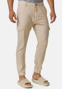 Beige linen cargo pants with tapered fit, elastic ankle cuffs, front and side pockets, and a button fly closure. Worn with beige slide sandals.