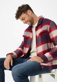 Red and cream checked flannel jacket with blue accents and two chest pockets, layered over a cream knitted sweater, paired with blue jeans.