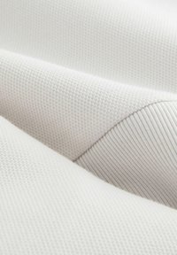 Tissu blanc texturé avec une surface côtelée, présentant une apparence tissée douce. Le matériau est lisse, formant des plis délicats.
