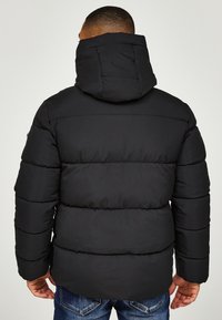 Svart puffjacka med horisontell quiltad design och huva, tillverkad av en slät tissu. Har en dragkedja och en rymlig passform, kombinerad med denimjeans.