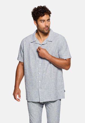 Hombre con cabello rizado que lleva una camisa de manga corta gris claro con botones y pantalones a juego, ajustándose el cuello de la camisa, mirando hacia un lado.