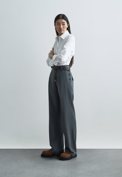 Chemise blanche à boutons avec des détails froncés, pantalons larges gris avec des boutons sur les côtés, et chaussures plateformes marron avec des semelles noires.