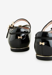 Zapatos de charol negros con punta redonda y un pequeño detalle de moño dorado, con una correa en el tobillo y un acento dorado en la suela.