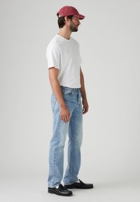 Camiseta de algodón blanca, jeans de mezclilla azul claro, gorra roja, mocasines negros. El modelo se sostiene de lado, mostrando un corte relajado y un estilo casual.