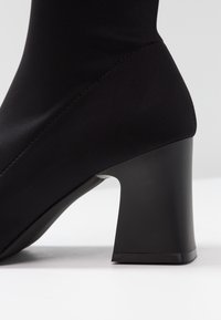 Bottine en tissu noir au design épuré, talon carré, texture lisse et détails cousus sur le côté.