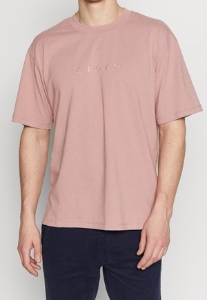 Mann trägt ein pinkes T-Shirt mit kurzen Ärmeln und japanischen Schriftzeichen auf der Brust sowie dunkle Hosen, steht vor einem einfarbigen Hintergrund.