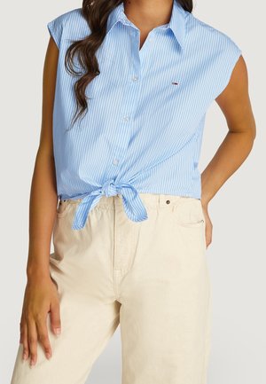 Light blue, sleeveless button-up shirt with white stripes, knotted at the waist, paired with cream-colored pants. Subtle logo detail present.