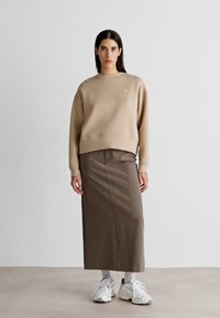 Sudadera beige con puños acanalados, combinada con una falda marrón hasta la rodilla. Llevada con zapatillas blancas y calcetines de color claro. Fondo liso.