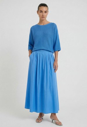 Femme debout les mains dans les poches, vêtue d’un haut en maille bleu ample, d’une longue jupe plissée bleue et de sandales argentées à talons.