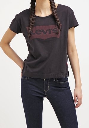 Kobieta z zaplecionymi włosami, ubrana w czarny t-shirt Levi's i ciemnoniebieskie dżinsy, pozująca z jedną ręką na biodrze.