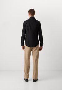 Zwart langemouwen shirt met een getailleerde pasvorm, knoopmanschetten en een gebogen zoom; gecombineerd met beige broek en zwarte schoenen.