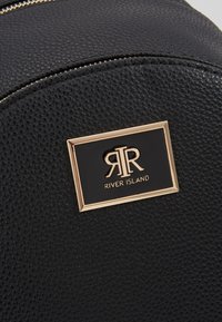 Czarny plecak z teksturowanej skóry z złotą, prostokątną plakietką logo River Island z przodu. Zamek błyskawiczny u góry.