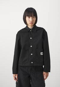 Veste en coton noire avec un col, fermeture à boutons à l'avant, deux poches et une étiquette de marque en bas à gauche.