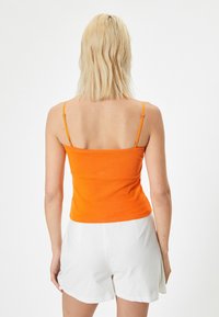 Orangefärgad anpassad linne med justerbara axelremmar, enkel design, texturerad bomullstyg, parat med vita shorts. Visas bakvy.