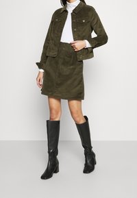 Olive green corduroy jacket with matching skirt, paired with black knee-high heeled boots. Features button details and a textured finish.
