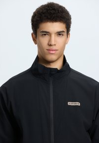 Black jacket with high collar, featuring a front zipper and a small logo on the left chest. Fabric appears smooth with a matte finish.