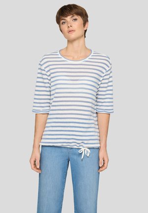Frau trägt ein hellblau-weiß gestreiftes Kurzarmshirt mit Kordelzug am Saum und hellblaue Jeans vor schlichtem Hintergrund.