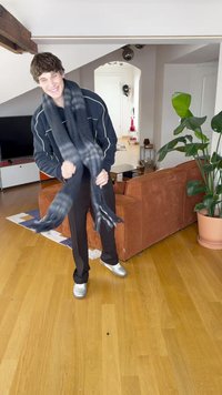 Μαύρο και γκρι καρό κασκόλ που φοριέται από ένα άτομο, φτιαγμένο από μαλακό ύφασμα με κρόσσια στις άκρες, πάνω σε ξύλινο πάτωμα και φόντο έναν καφέ καναπέ.