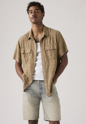 Homme aux cheveux courts et bouclés, portant une chemise beige à manches courtes avec imprimé floral par-dessus un débardeur blanc et un short en jean délavé, debout les mains dans les poches.