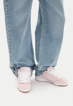 Jasnoniebieskie szerokie jeansy zestawione z pastelowo różowymi zamszowymi trampkami na białym tle, z białymi sznurówkami.