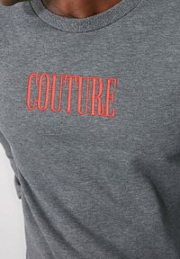 Sudadera gris con cuello acanalado, que presenta la palabra "COUTURE" en letras grandes y de un rojo brillante en el pecho. Material de algodón suave.