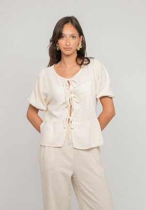 Femme aux longs cheveux noirs portant un chemisier crème texturé à manches courtes avec trois nœuds noués et un pantalon ample assorti, debout les mains derrière le dos.