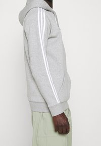 Persona con sudadera con capucha gris claro y rayas blancas en las mangas, y pantalones verdes claros, mostrada de lado.