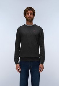 Man with curly hair and beard wearing a dark gray long-sleeve sweater and blue jeans standing against a plain light background.