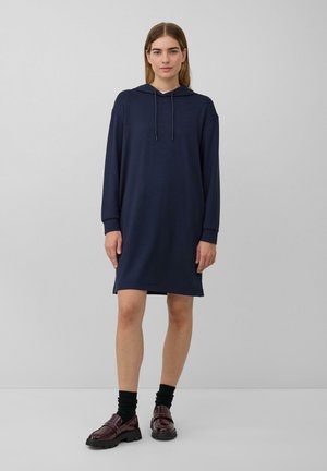 Vrouw staat in een marineblauw langslavend hoodie-jurk, zwarte sokken en donker bordeauxrode grove instappers tegen een effen lichte achtergrond.