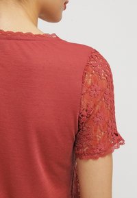 Roodachtig-pink shirt met korte kantensleeves, voorzien van bloemenpatronen en golvende randen. Glad katoenen lichaam met transparante kantdetails.