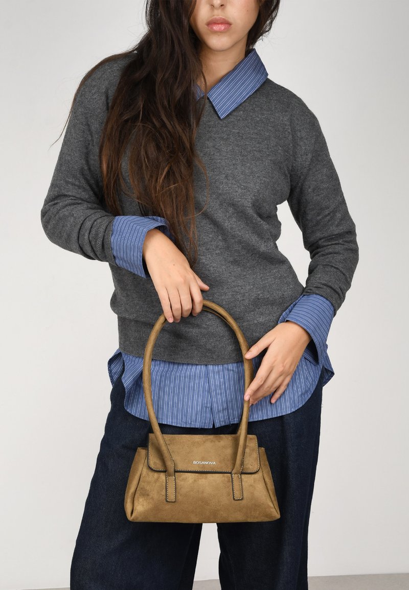 Bolso de piel ante color beige con un diseño estructurado y asas cortas. Lleva un suéter gris sobre una camisa de rayas azul, combinado con jeans oscuros.