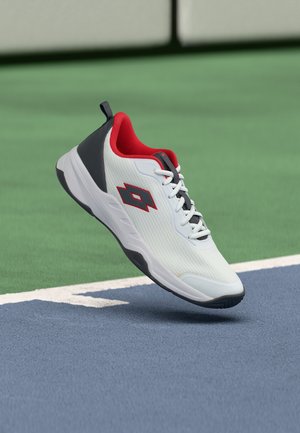 Valge spordijalats mustade ja punaste detailidega, hõljumas sinise tennisekatuse kohal valge piirjoone lähedal.