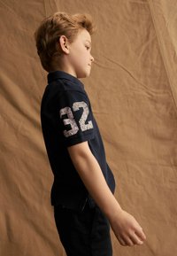 Jūrinis polo marškinėliai su baltu tekstūruotu "32" aplikacijomis ant rankovės, trumpomis rankovėmis ir plokščiu apykakle, derinami su tamsiomis kelnėmis. Lygus audinio paviršius.