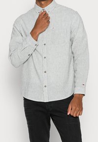 Camisa de botones a rayas verdes y blancas en tejido de algodón, con mangas largas, bolsillo en el pecho y botones plateados. Acentuada con un parche de logo.