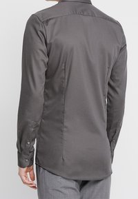 Szara koszula z długim rękawem o gładkiej fakturze, z mankietem na guziki, zaszewką na plecach i klasycznym kołnierzykiem. Fason dopasowany, z zaokrąglonym dołem.