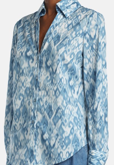 Tričko s dlhým rukávom na gombíky v svetlomodrej farbe s abstraktným ikatovým vzorom. Vyrobené z mäkkej látky, má golier a zaoblený lem.