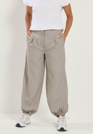 Personne portant un pantalon beige à fines rayures fuselé avec des poignets élastiques, des baskets blanches et un haut blanc uni à manches courtes, mains dans les poches.